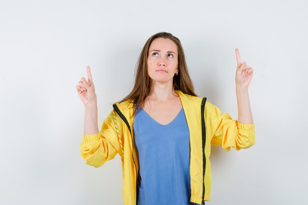 Young lady pointing up in t-shirt, jacket and looking pensive, front view.