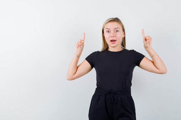 Free photo young lady pointing up in t-shirt, pants and looking merry. front view.