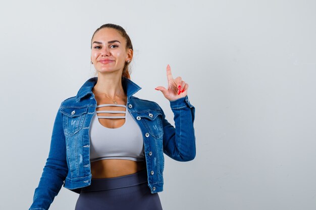 Young lady pointing up in top, denim jacket and looking pleased , front view.