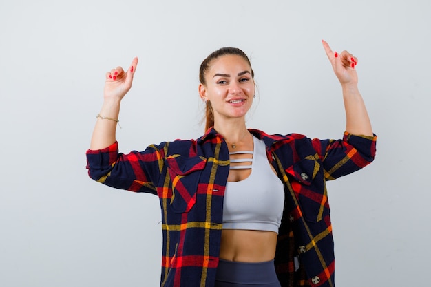 Young lady pointing up in top, plaid shirt and looking cheery , front view.