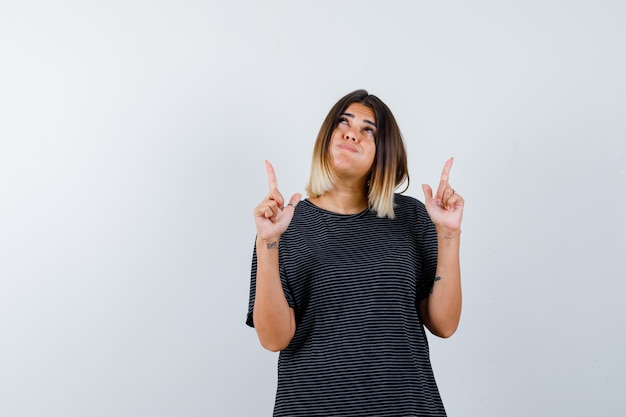 Young lady pointing up while blowing cheeks in polo dress and looking hesitant , front view.