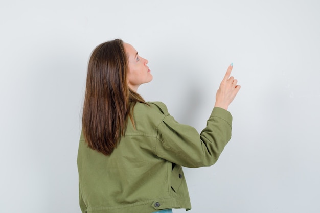 Young lady pointing up while looking upward in green jacket and looking focused , back view.