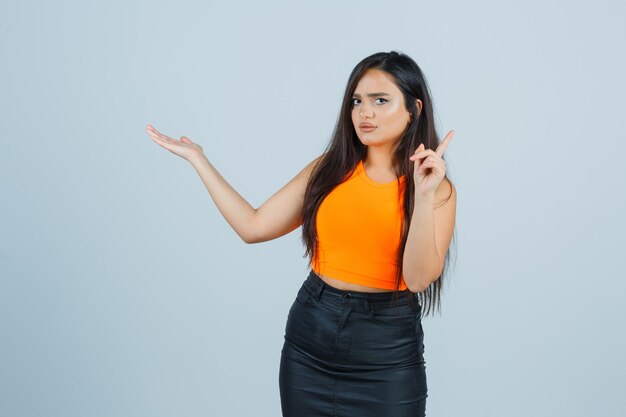 Young lady pointing up while presenting something in singlet, mini skirt and looking puzzled , front view.