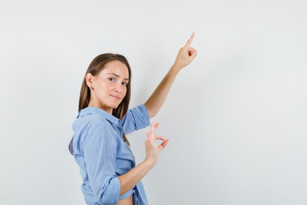 Young lady pointing up while showing ok sign in blue shirt and looking pleased.