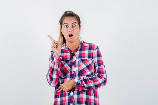 Young lady pointing at upper left corner in checked shirt and looking surprised , front view.