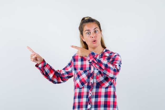 Young lady pointing at upper left corner in checked shirt and looking wondered , front view.