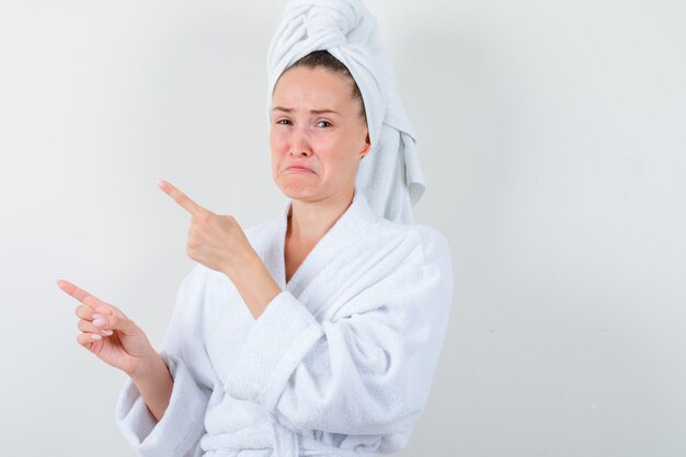 Юная леди, указывая на верхний левый угол в белом халате, полотенце и разочарованно, вид спереди.