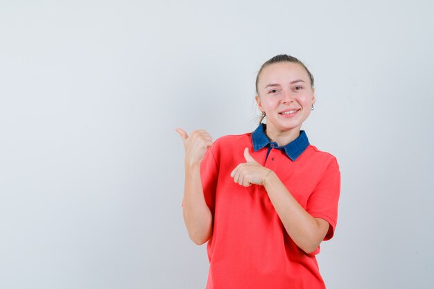 Young lady pointing at upper left corner with thumbs in t-shirt and looking glad