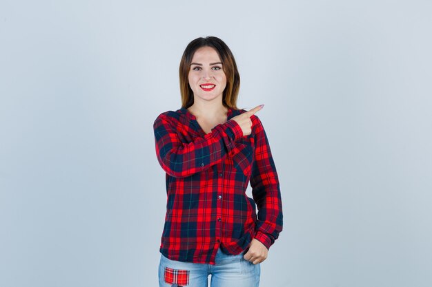 Young lady pointing at upper right corner in checked shirt and looking cheerful , front view.