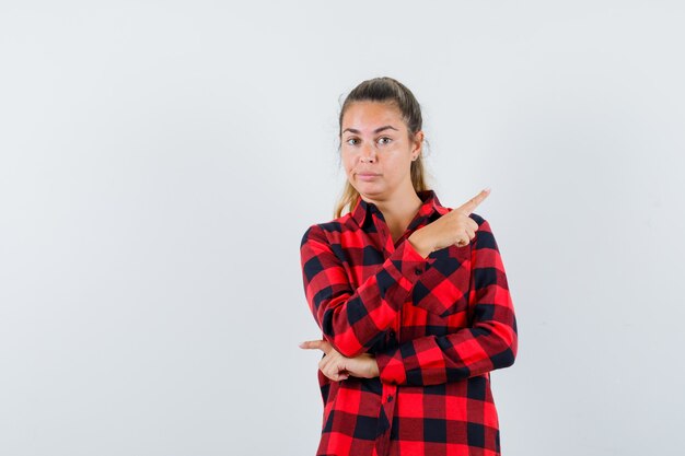 Young lady pointing at upper right corner in checked shirt and looking hesitant