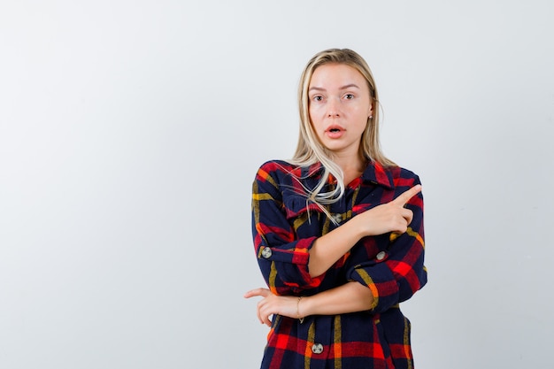 Young lady pointing at upper right corner in checked shirt and looking puzzled , front view.