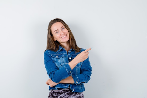 Young lady pointing at upper right corner in denim jacket and looking merry , front view.