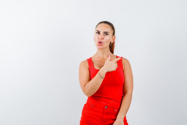 Young lady pointing at upper right corner in red singlet, red trousers and looking perplexed , front view.