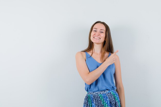 Young lady pointing at upper right corner in singlet, skirt and looking happy , front view.