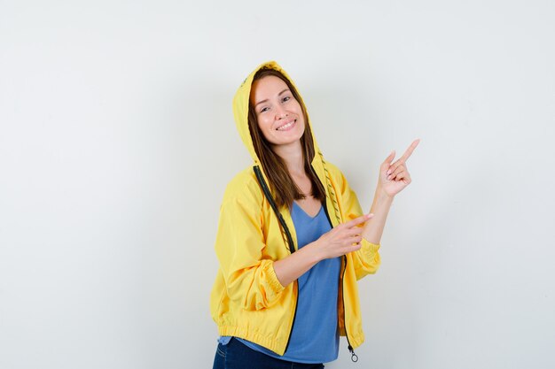 Young lady pointing at upper right corner in t-shirt, jacket and looking merry, front view.