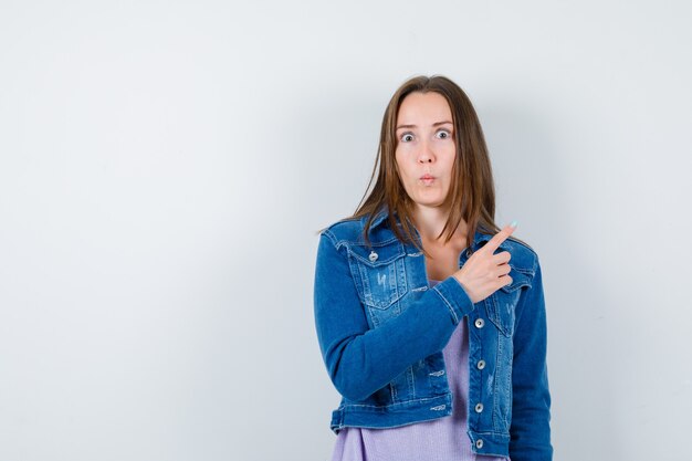 Young lady pointing at upper right corner in t-shirt, jacket and looking scared , front view.