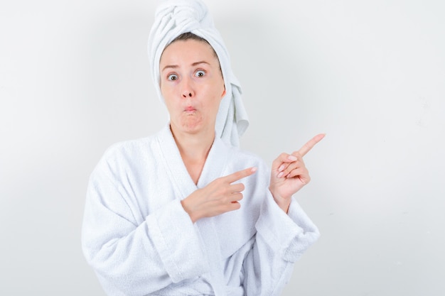 Young lady pointing at upper right corner in white bathrobe, towel and looking amazed. front view.