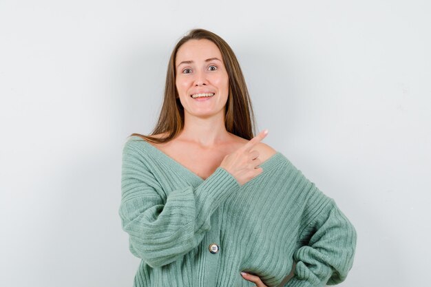 Young lady pointing at upper right corner in wool cardigan and looking happy. front view.