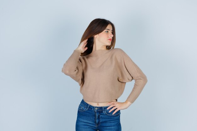 Young lady posing while arranging her hear in beige sweater, jeans and looking peaceful. front view.