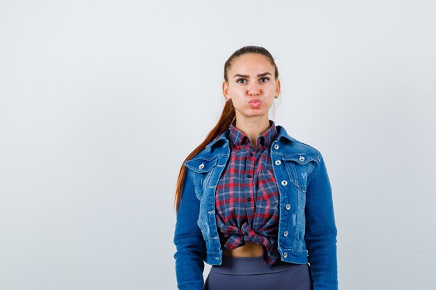Young lady posing while looking, blowing cheeks in checkered shirt, denim jacket and looking funny , front view.