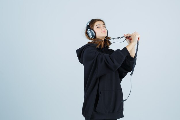 Young lady posing while pulling handphones in oversized hoodie, pants and looking funny , front view.