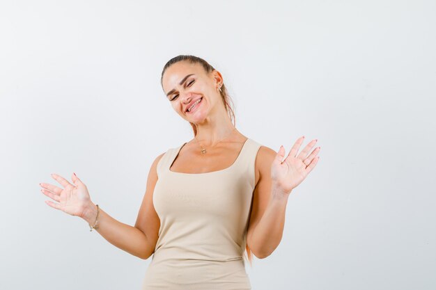 Young lady posing while showing palms in beige tank top and looking cheerful , front view.