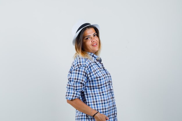 Young lady posing while standing in checked shirt hat and looking confident 
