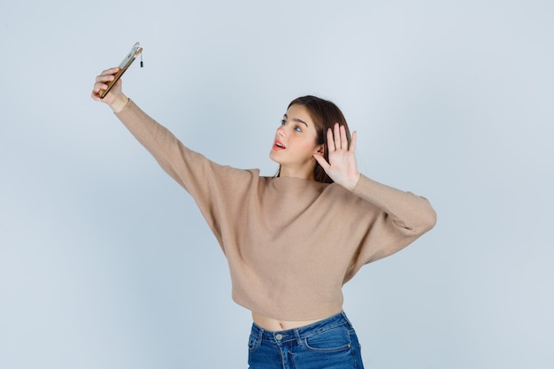 Giovane donna in posa mentre si fa selfie con il telefono cellulare, agitando la mano in maglione beige, jeans e guardando attraente, vista frontale.