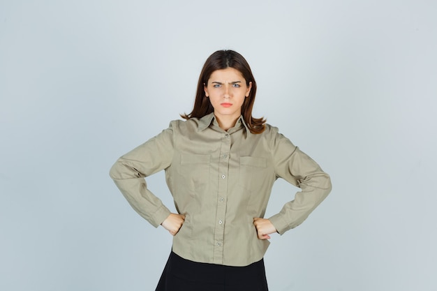 Free photo young lady posing with fists on waist in shirt, skirt and looking annoyed