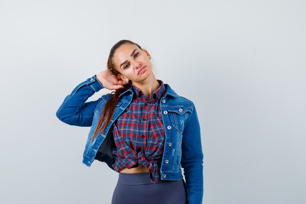 Young lady posing with hand behind head in checkered shirt, denim jacket and looking confident , front view.