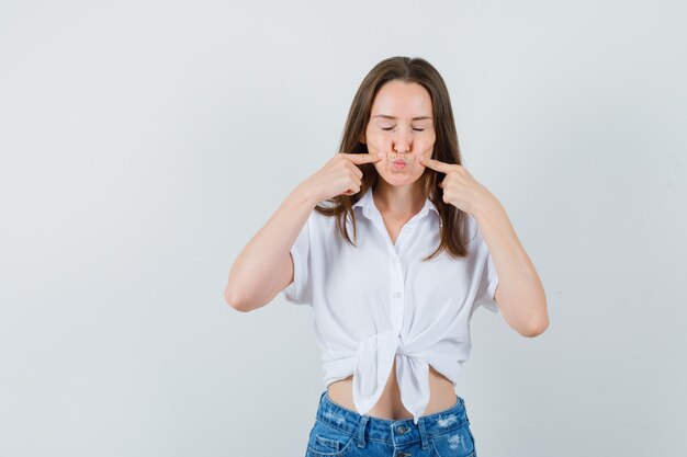 Young lady pouting her lips while squeezing cheeks with fingers in white blouse , front view.