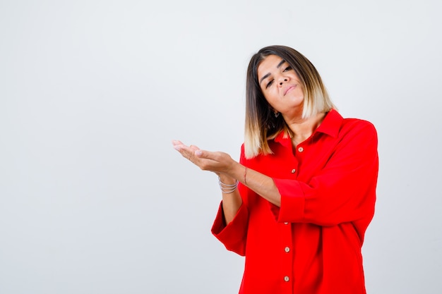 Young lady pretending to hold or show something in red oversize shirt and looking confident. front view.