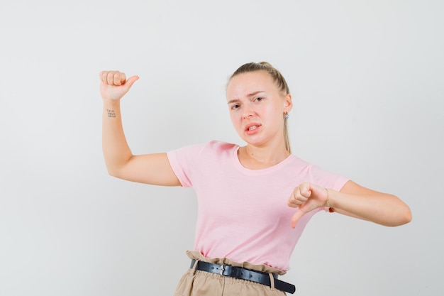 Young lady pretending to hold something, showing thumb down in t-shirt and pants and looking dissatisfied, front view