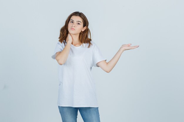 Young lady pretending to show something while holding hand on neck in t-shirt, jeans and looking confident , front view.