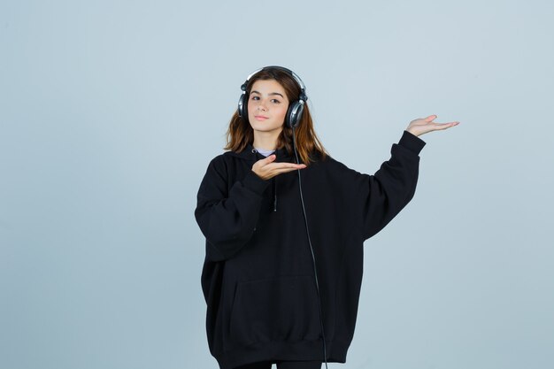 Young lady pretending to show something while listening to music with headphones in oversized hoodie, pants and looking confident. front view.