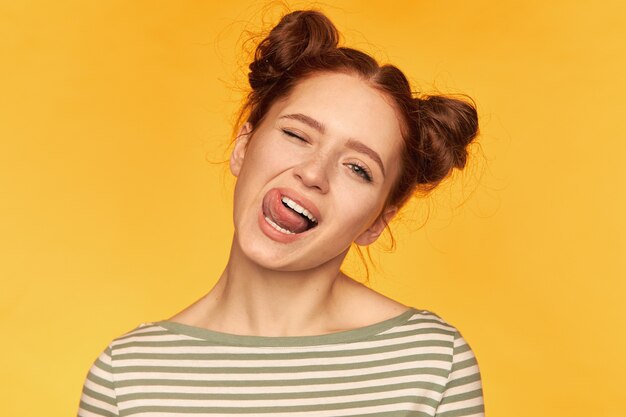 Young lady, pretty ginger woman with two buns. have a playful, silly mood. wearing striped sweater, wink and show a tongue isolated, closeup over yellow wall