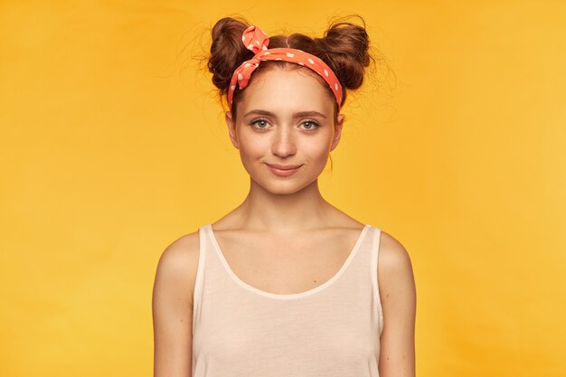Free photo young lady, pretty ginger woman with two buns. wearing white tank top and red doted hairband. looking confident, waiting for something.  isolated over yellow wall