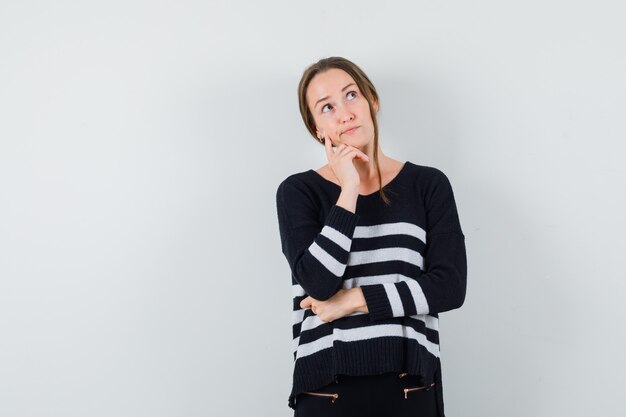 Young lady propping chin on hand in casual shirt and looking hesitant