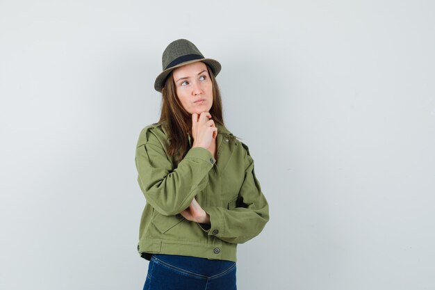 Young lady propping chin on hand in jacket pants hat and looking pensive 