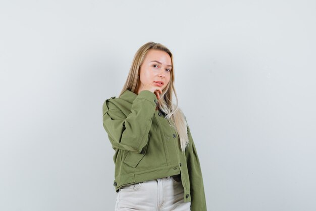 Young lady propping chin on hand in jacket, pants and looking delicate , front view.
