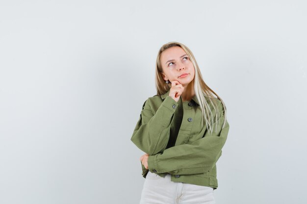 Young lady propping chin on hand in jacket, pants and looking dreamy. front view.