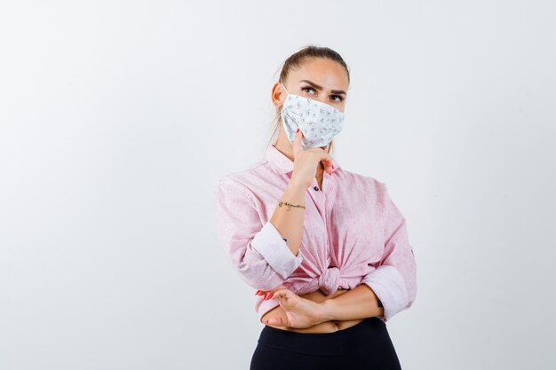 Free photo young lady propping chin on hand in shirt, mask and looking pensive. front view.