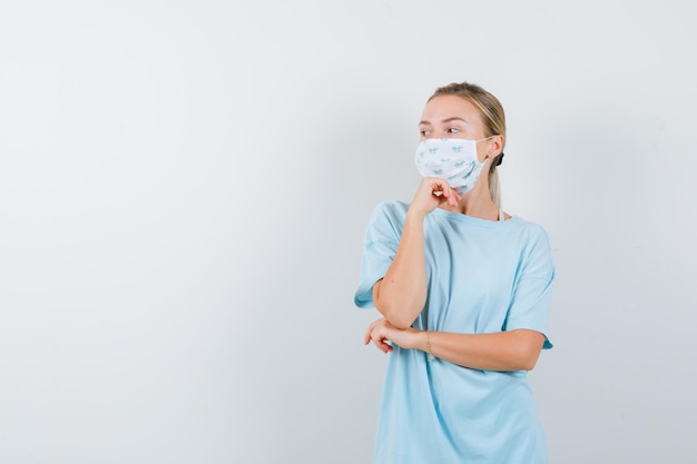 Young lady propping chin on hand in t-shirt, mask and looking pensive