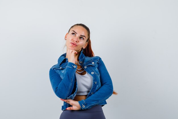 Young lady propping chin on hand in top, denim jacket and looking pensive. front view.