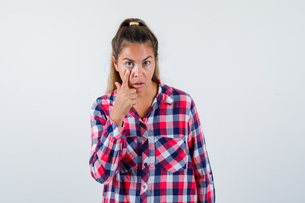 Young lady pulling down her eyelid in checked shirt and looking puzzled. front view.