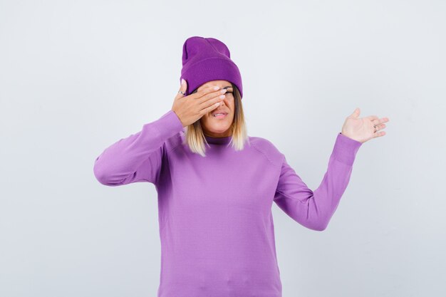紫色のセーターを着た若い女性、何かを見せて陽気に見えながら、目の上に手を置いたビーニー、正面図。