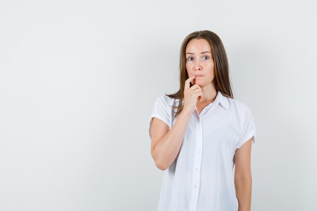 Young lady putting finger on her mouth in white blouse and looking thoughtful