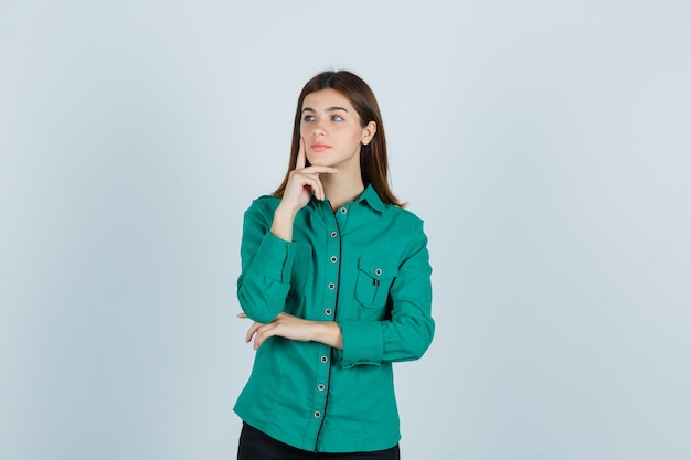 緑のシャツのあごを支えるために指を置き、物思いにふける、正面図を探している若い女性。