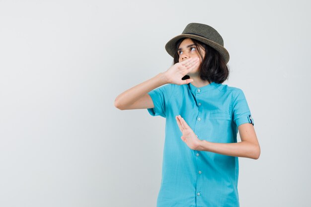 Young lady raising hand to defend herself in blue shirt, hat and looking concentrated.