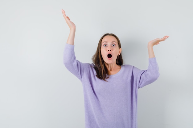 Young lady raising hands in casual shirt and looking surprised. front view.